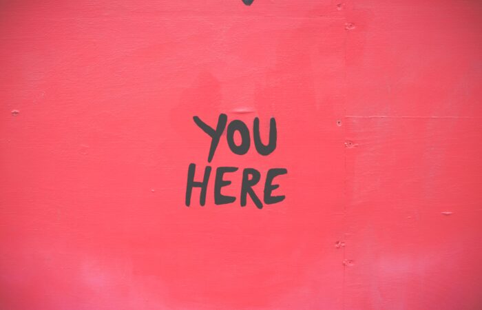 Φράση “YOU HERE” γραμμένη με μαύρα γράμματα σε ροζ τοίχο – minimal εικόνα που υποδηλώνει παρουσία και θέση.
