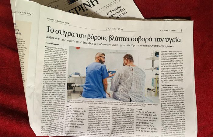 Ανοιχτό φύλλο της εφημερίδας 'ΤΟ ΘΕΜΑ' ακουμπισμένο σε κόκκινο ύφασμα. Η σελίδα που διακρίνεται έχει τίτλο 'Το στίγμα του βάρους βλάπτει σοβαρά την υγεία' από τη δημοσιογράφο Λίνα Γιάνναρου. Ο υπότιτλος αναφέρει: 'Άνθρωποι με παχυσαρκία συχνά διστάζουν να αναζητήσουν ιατρική φροντίδα λόγω διακρίσεων που έχουν βιώσει'. Στη μέση του άρθρου, υπάρχει φωτογραφία ενός γιατρού με μπλε στολή που συζητά με έναν άνδρα με παχυσαρκία, ο οποίος φοράει νοσοκομειακή ρόμπα.